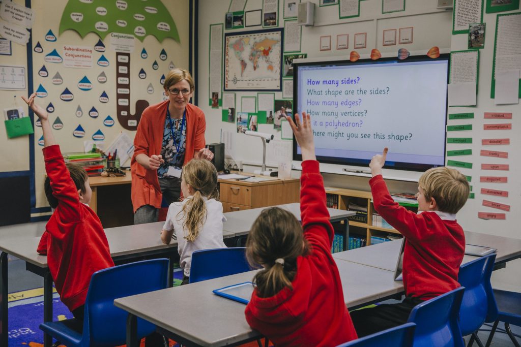 Female teacher is teaching shapes to her primary school students. She is asking hem a question and some of the students have their hand in the air to answer.